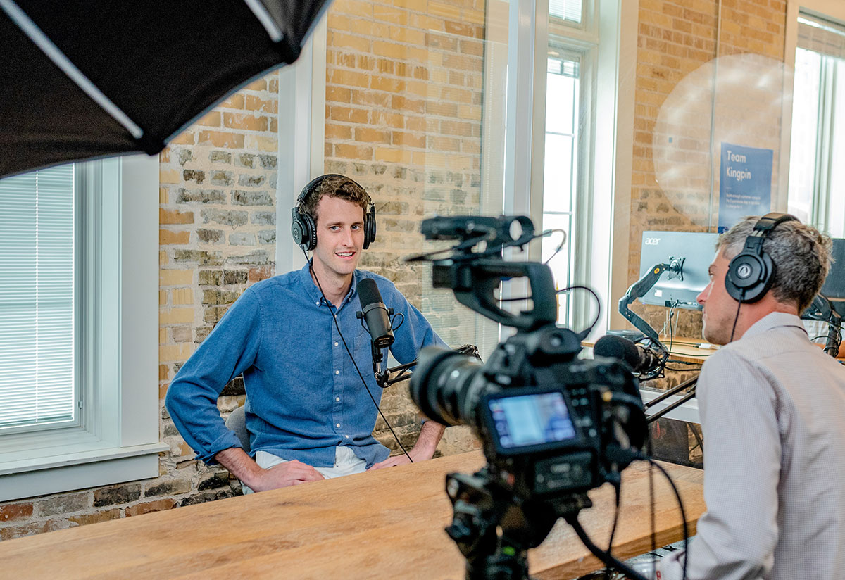 Two men sit in a modern offices with big headphones on. One is recording a podcast with a microphone in front of him. The other is sitting next to a camera.