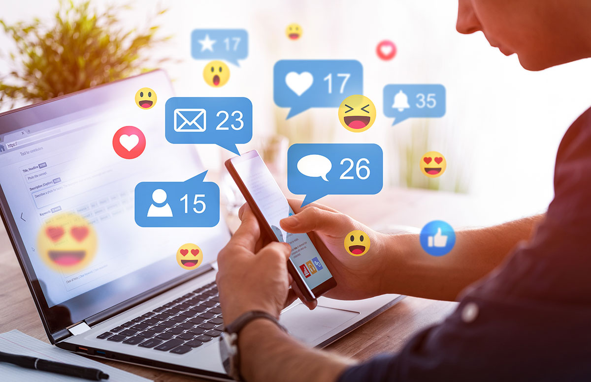 A man is looking at his phone with his computer behind it. There are social media icons popping up, including emojis, like notifications and messenger notifications. 