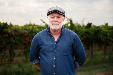 A man with a silver beard wearing a denim shirt and flat cap poses in front of a vineyard