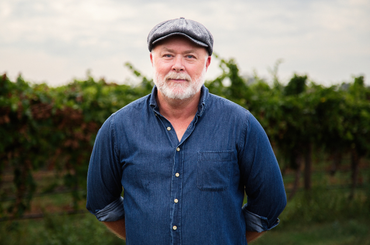 A man with a silver beard poses in a vineyard