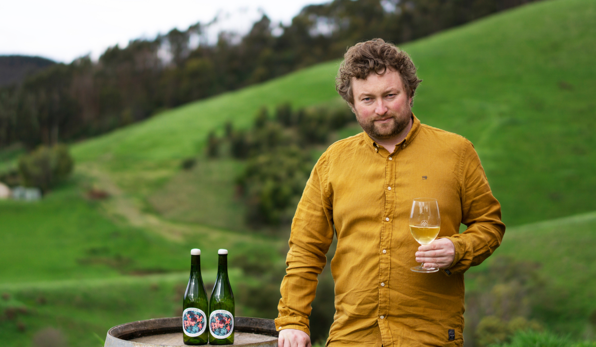 A man stands next to a wine barrel holding a glass of wine. Two bottles of wine are on the barrel.