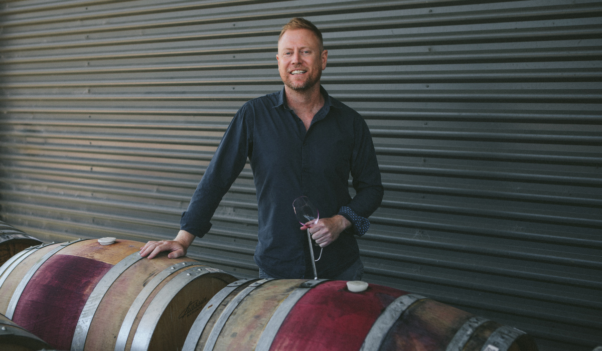 A winemaker stands behind a wine barrel holding a wine glass