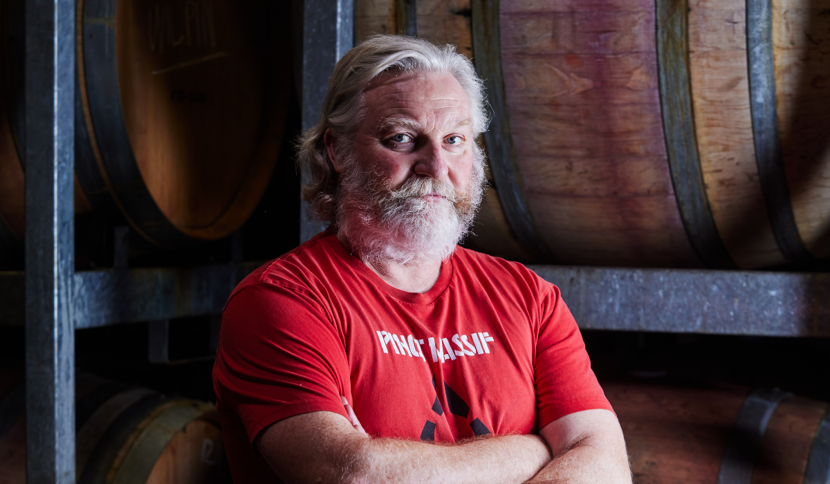 A man wearing a red shirt crosses his arms in front of wine barrels