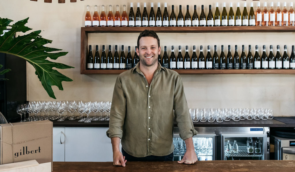 A man stands behind a bar, bottles of wine are on display behind him