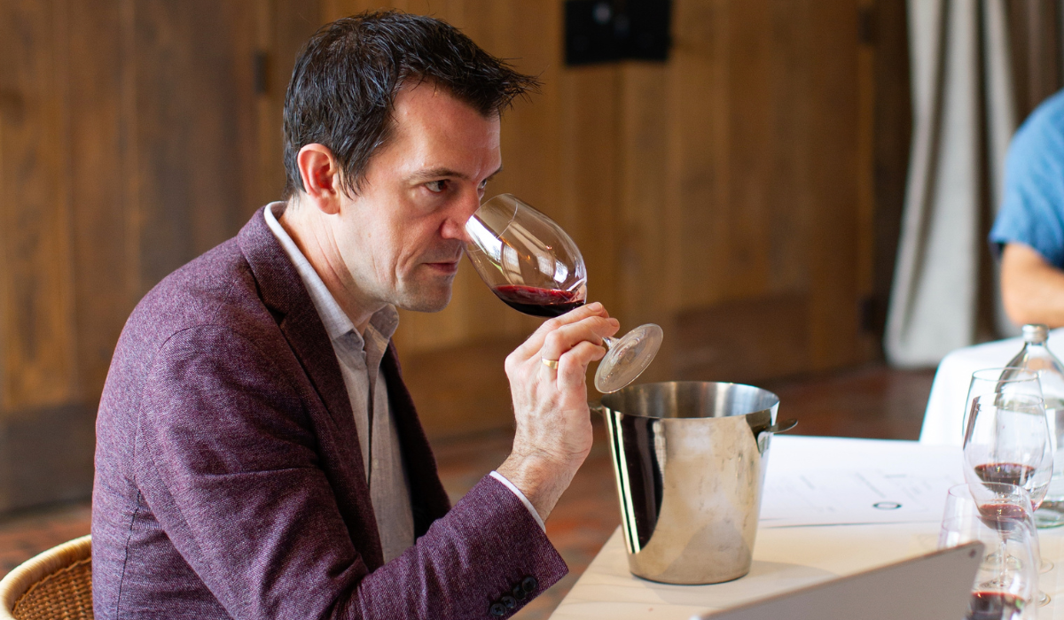 A man wearing a purple blazer sniffs a glass of red wine