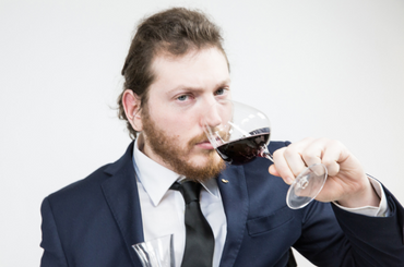 A man with a beard and a ponytail smells a glass of red wine and stares into the camera