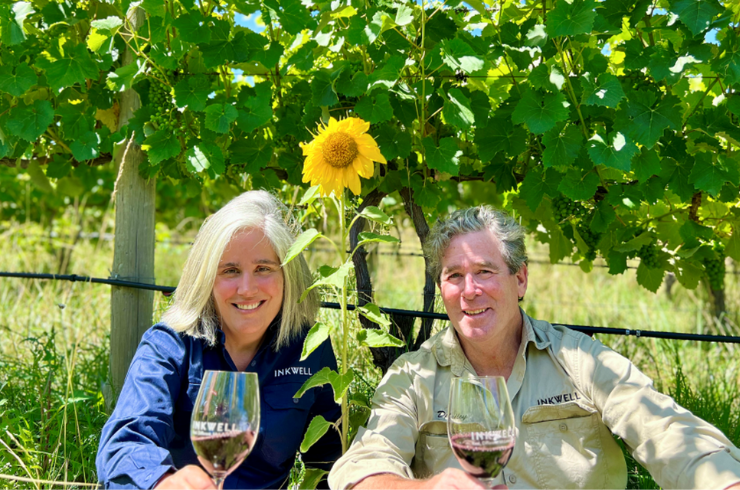 A woman and a man pose in front of a vineyard with a sunflower growing in it