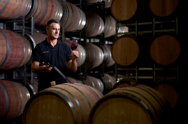 A winemaker stands behind some wine barrels, siphoning a taste