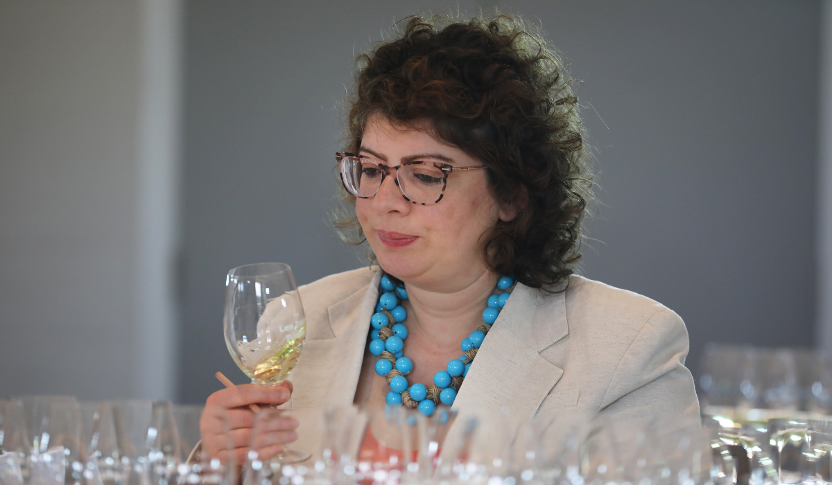 A woman with curly brown hair tastes judges white wine