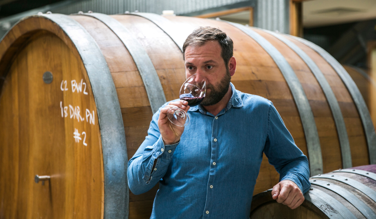 A winemaker tastes a glass of red wine in front of a barrel