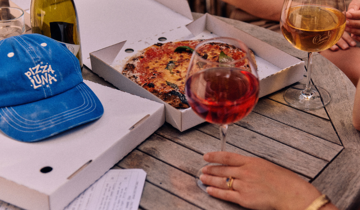 A blue cap, pizza and wine on a table