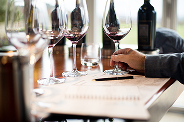 A man's hand holding a glass of red wine. There are wine bottles and wine glasses in the background, and a notepad and pen in front.