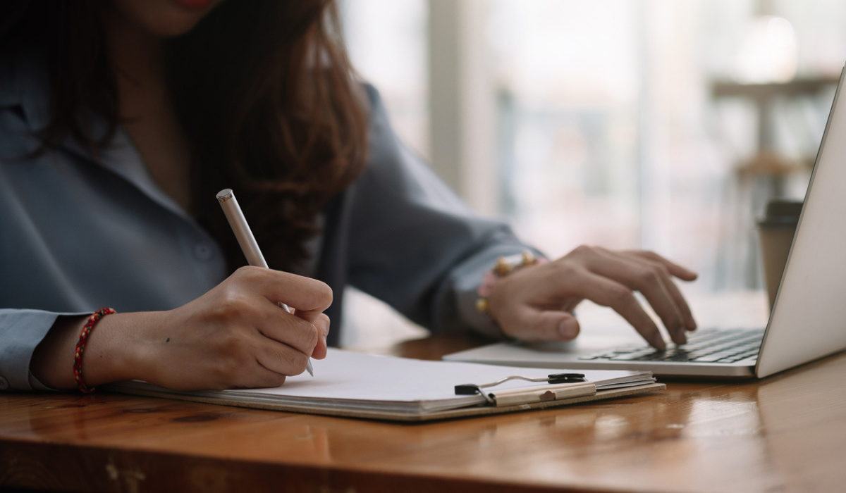 Person writing notes on paper while using a laptop